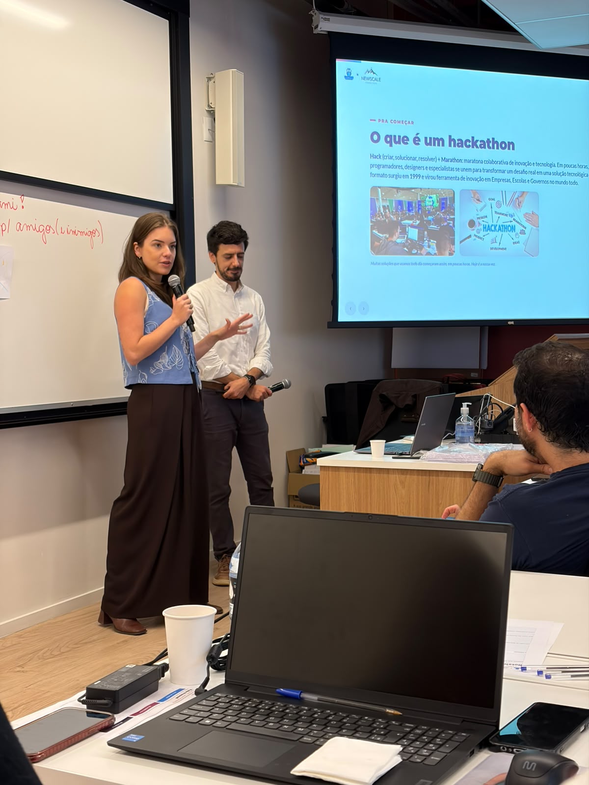 Bruna Santanna conduzindo a abertura do hackathon de IA no palco, com o slide O que é um hackathon